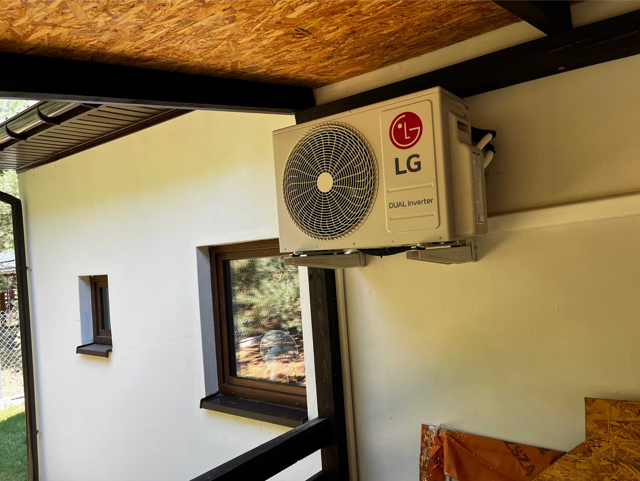 Zewnętrzna jednostka klimatyzatora LG Dual Inverter zamontowana na ścianie budynku z widocznymi oknami i drewnianym zadaszeniem.