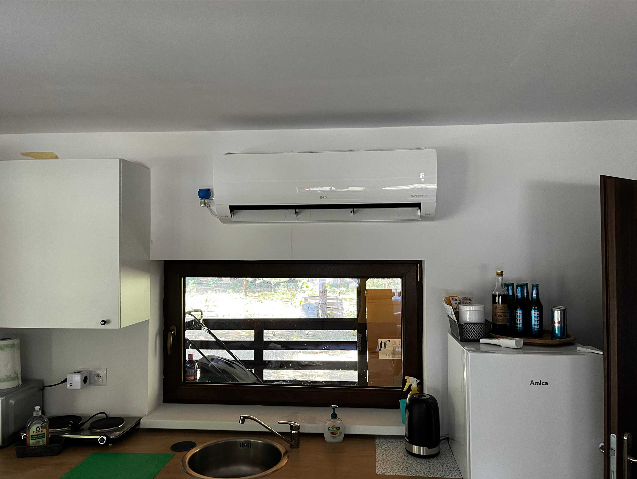 Biały klimatyzator LG zamontowany na ścianie w kuchni nad oknem, z widocznym wyświetlaczem temperatury i niebieskim zaworem na boku, otoczenie kuchenne z szafkami, zlewem, i lodówką Amica.