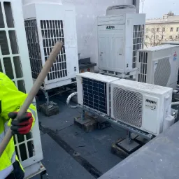 Pracownik w odblaskowej kurtce i rękawicach roboczych z długim kijem na dachu pokrytym papą, w tle liczne jednostki klimatyzacyjne różnych marek (Aermec, Mitsubishi) zamontowane na betonowych...