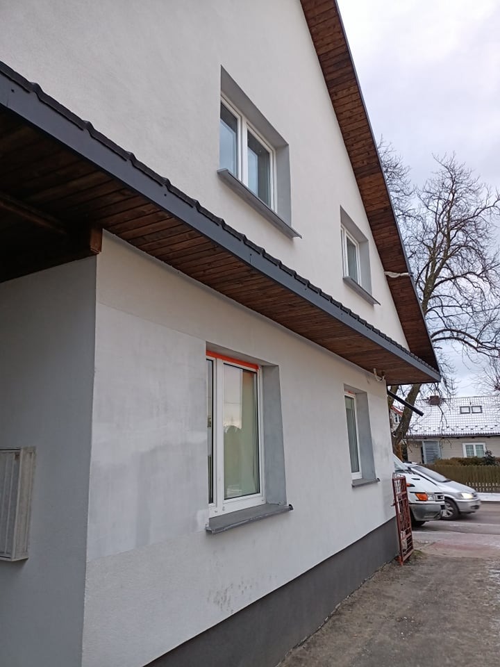 Boczny widok odnowionej elewacji dwukondygnacyjnego domu z białymi ścianami i ciemnym drewnianym podbitką dachową, widoczne okna z białymi ramami i szary cokół, na tle pochmurne niebo i drzewo bez...