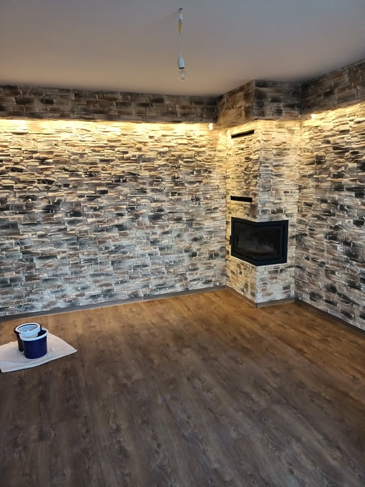 Wnętrze pokoju z kamienną okładziną ścian i kominkiem narożnym, podświetlenie LED przy suficie, ciemna podłoga z paneli, dwa wiadra na podłodze.