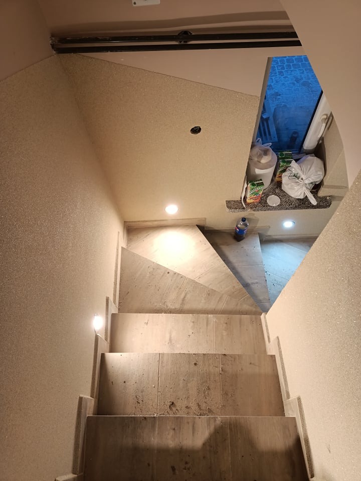 Widok z góry na klatkę schodową w trakcie prac wykończeniowych, z widocznymi schodami z jasnych płytek, oświetleniem punktowym w ścianach i suficie, oraz materiałami budowlanymi przy oknie.