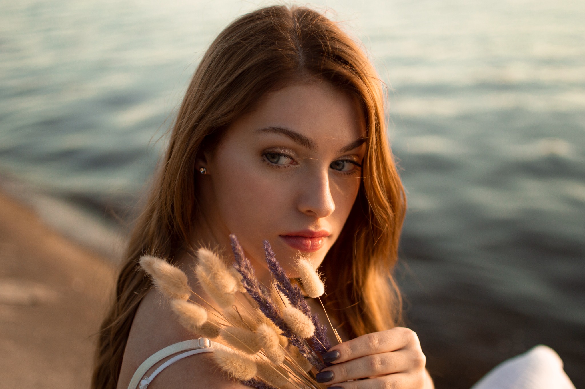 Portret młodej kobiety z rudymi włosami, trzymającej pęczek suszonych traw w odcieniach beżu i fioletu na tle rozmytej wody o zachodzie słońca.