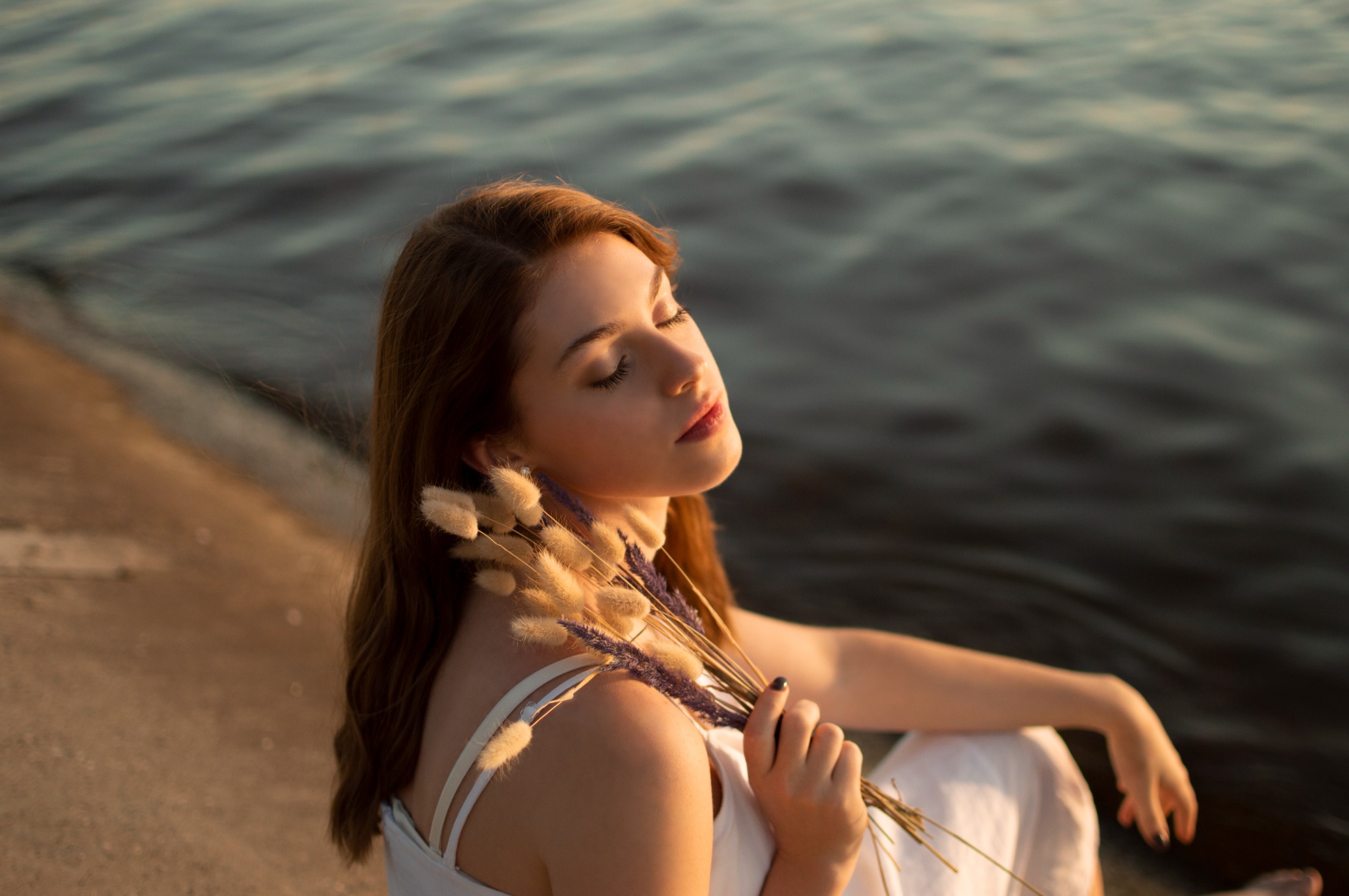 Młoda kobieta w białej sukience siedzi na brzegu zbiornika wodnego, trzymając bukiet suszonych traw w ciepłym, zachodzącym słońcu.