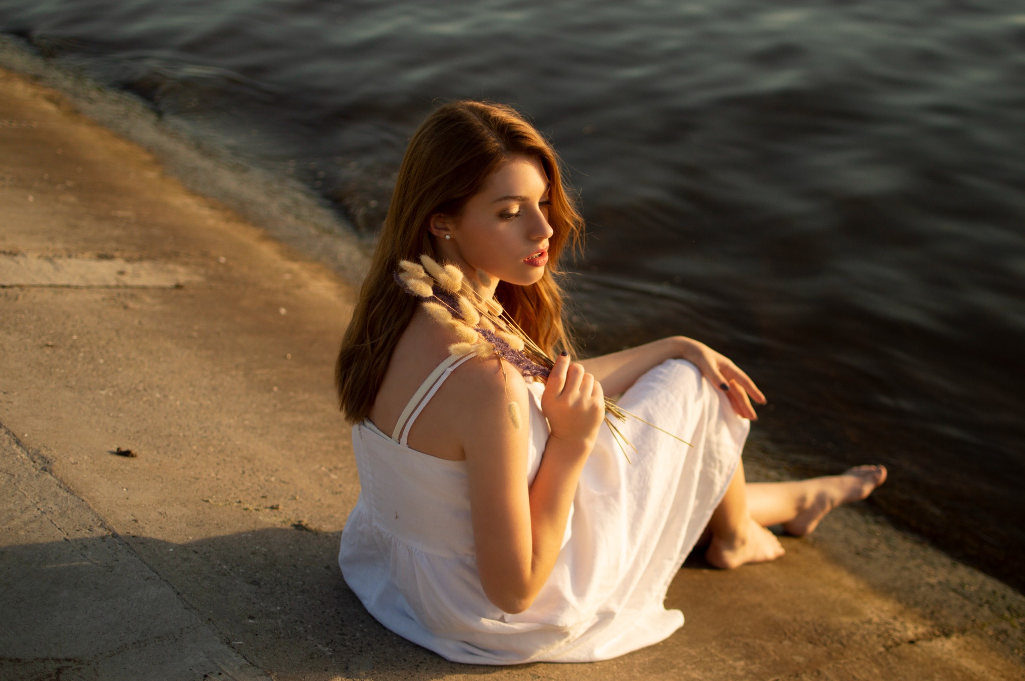 Młoda kobieta w białej sukience siedzi na betonowym nabrzeżu, trzymając pęk suszonych traw, z widokiem na wodę w ciepłym, zachodzącym słońcu.