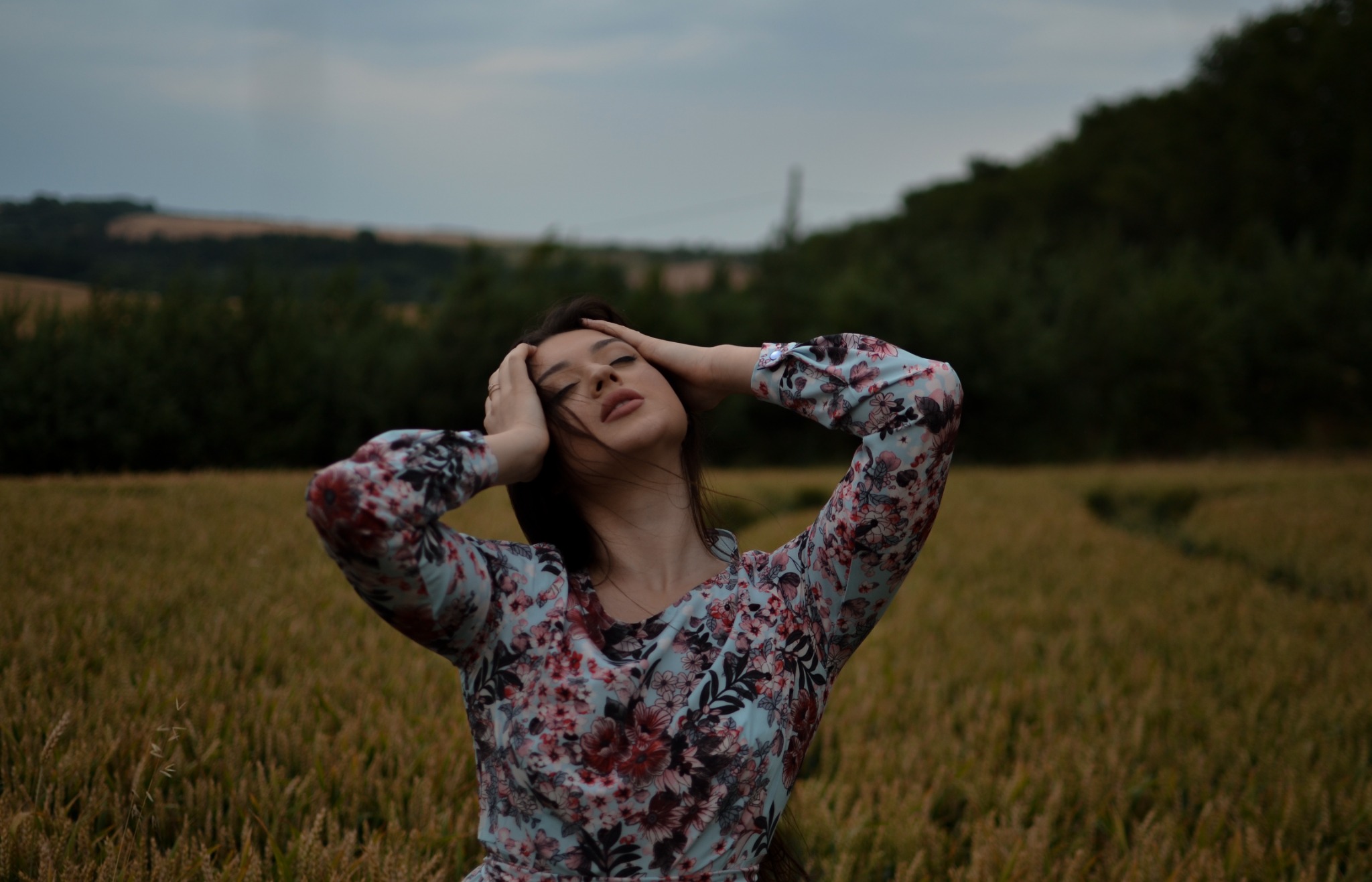Kobieta w kwiecistej sukience z zamkniętymi oczami, trzymająca ręce na głowie, stojąca na polu zboża z zalesionym wzgórzem w tle.