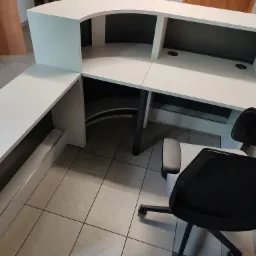 Nowoczesna, biała lada recepcyjna z czarnymi akcentami, widok z perspektywy, obok krzesło biurowe z siatkowym oparciem, na podłodze płytki ceramiczne.