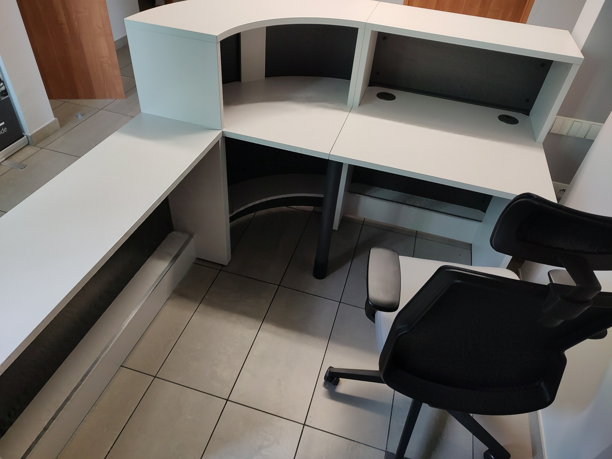 Nowoczesna, biała lada recepcyjna z czarnymi akcentami, widok z perspektywy, obok krzesło biurowe z siatkowym oparciem, na podłodze płytki ceramiczne.