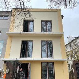 Olbud - Odnowiona elewacja trzypiętrowego budynku w odcieniach beżu i żółci, z ciemnymi oknami, widoczna brama wejściowa z pracownikiem w ubraniu roboczym i wiaderkiem, oraz fragment sąsiedniego budynku...