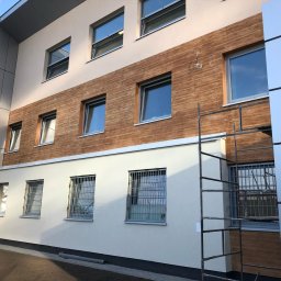 Olbud - Budynek po częściowym nałożeniu elewacji imitującej drewno, widoczne rusztowanie i okna z kratami.