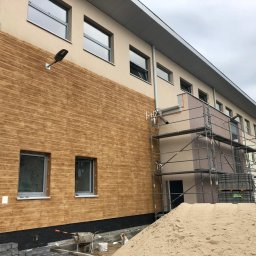 Olbud - Budynek z elewacją imitującą drewno, rusztowania, piasek, i materiały budowlane wskazują na prace wykończeniowe.