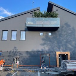 Olbud - Dwóch pracowników w kombinezonach maluje elewację budynku w kolorze grafitowym, z balkonem obsadzonym zielenią na górze; jeden z nich miesza farbę, drugi stoi obok drzwi; widoczny wózek z panelami.