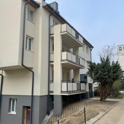 Olbud - Nowoczesny budynek wielorodzinny z jasną elewacją, szarym cokołem, balkonami z metalowymi balustradami i rynnami, widok z dołu na tle drzewa i sąsiedniego budynku.