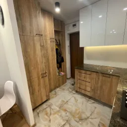 Wykończona kuchnia w Warszawie z drewnianymi szafkami i marmurową podłogą, widoczny fragment przedpokoju z wieszakami i szafką na buty.