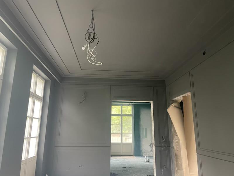 Wnętrze pokoju w trakcie remontu, świeżo pomalowane ściany w odcieniach szarości, widoczne wystające kable z żarówką na suficie, częściowo zasłonięte drzwi z roletą.