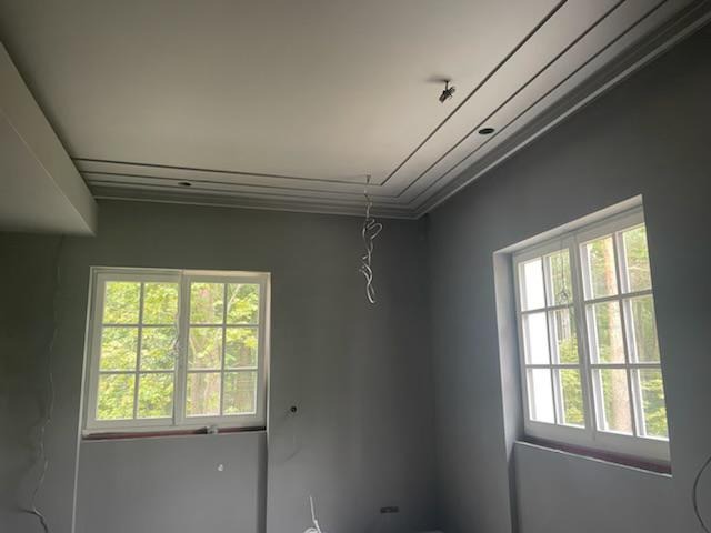 Szare wnętrze pokoju w trakcie remontu: widoczne dwa białe okna, surowa ściana i dekoracyjny sufit z listwami, zwisające kable