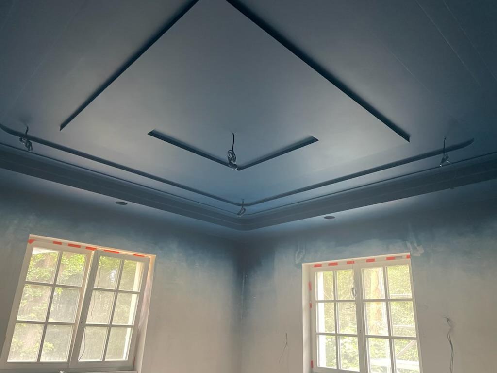 Niedokończone wnętrze pokoju z pomalowanym na niebiesko sufitem z dekoracyjnymi listwami, surowe ściany z widocznymi śladami malowania, dwa okna z widokiem na zieleń na zewnątrz i wystającymi...