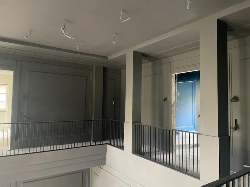 Wnętrze domu w trakcie remontu: szare ściany z panelami, czarne filary, metalowa balustrada, widoczne kable zwisające z sufitu i fragment niebieskiego pomieszczenia w tle, co wskazuje na trwające...