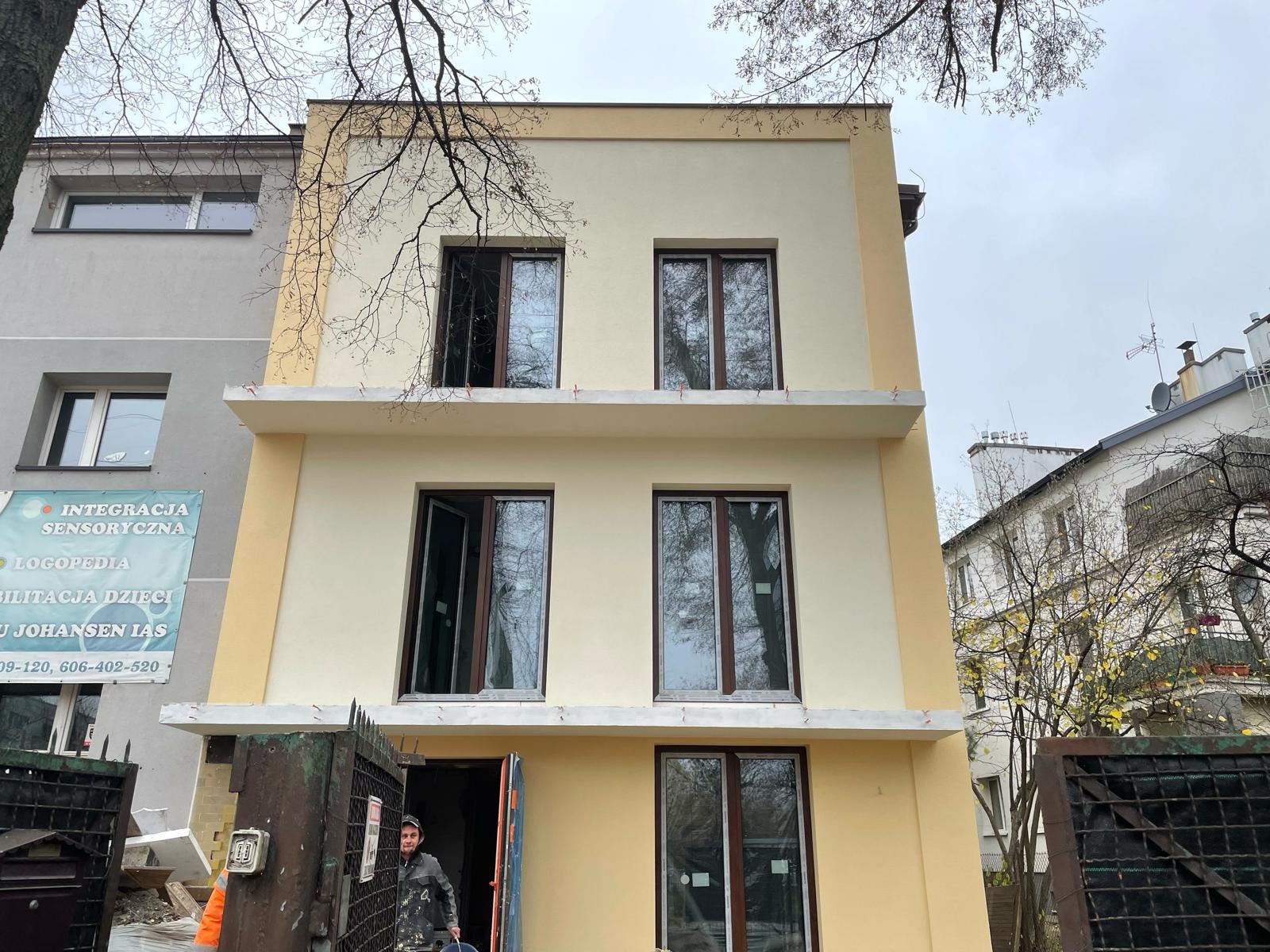 Odnowiona elewacja trzypiętrowego budynku w odcieniach beżu i żółci, z ciemnymi oknami, widoczna brama wejściowa z pracownikiem w ubraniu roboczym i wiaderkiem, oraz fragment sąsiedniego budynku...