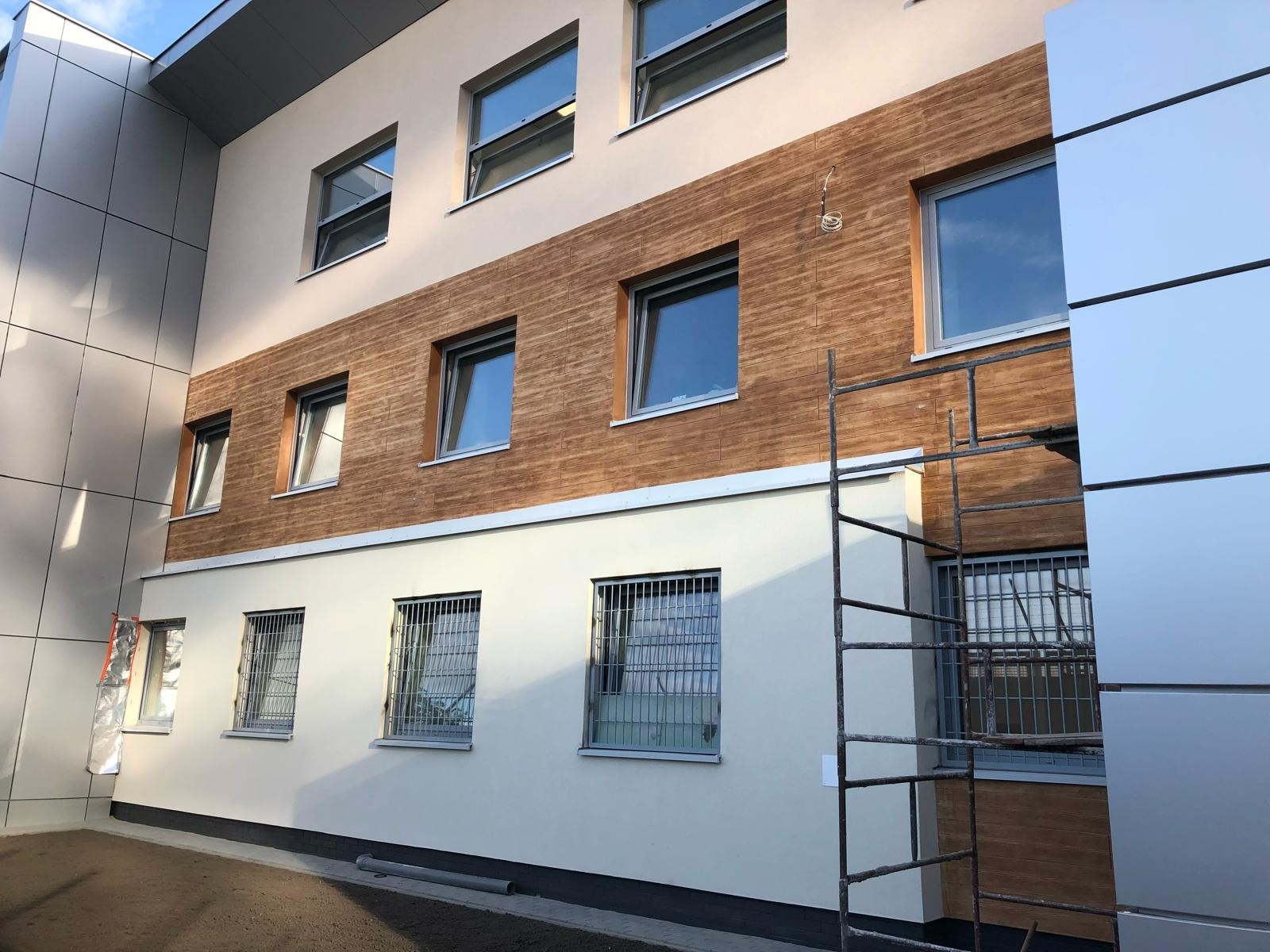 Budynek po częściowym nałożeniu elewacji imitującej drewno, widoczne rusztowanie i okna z kratami.