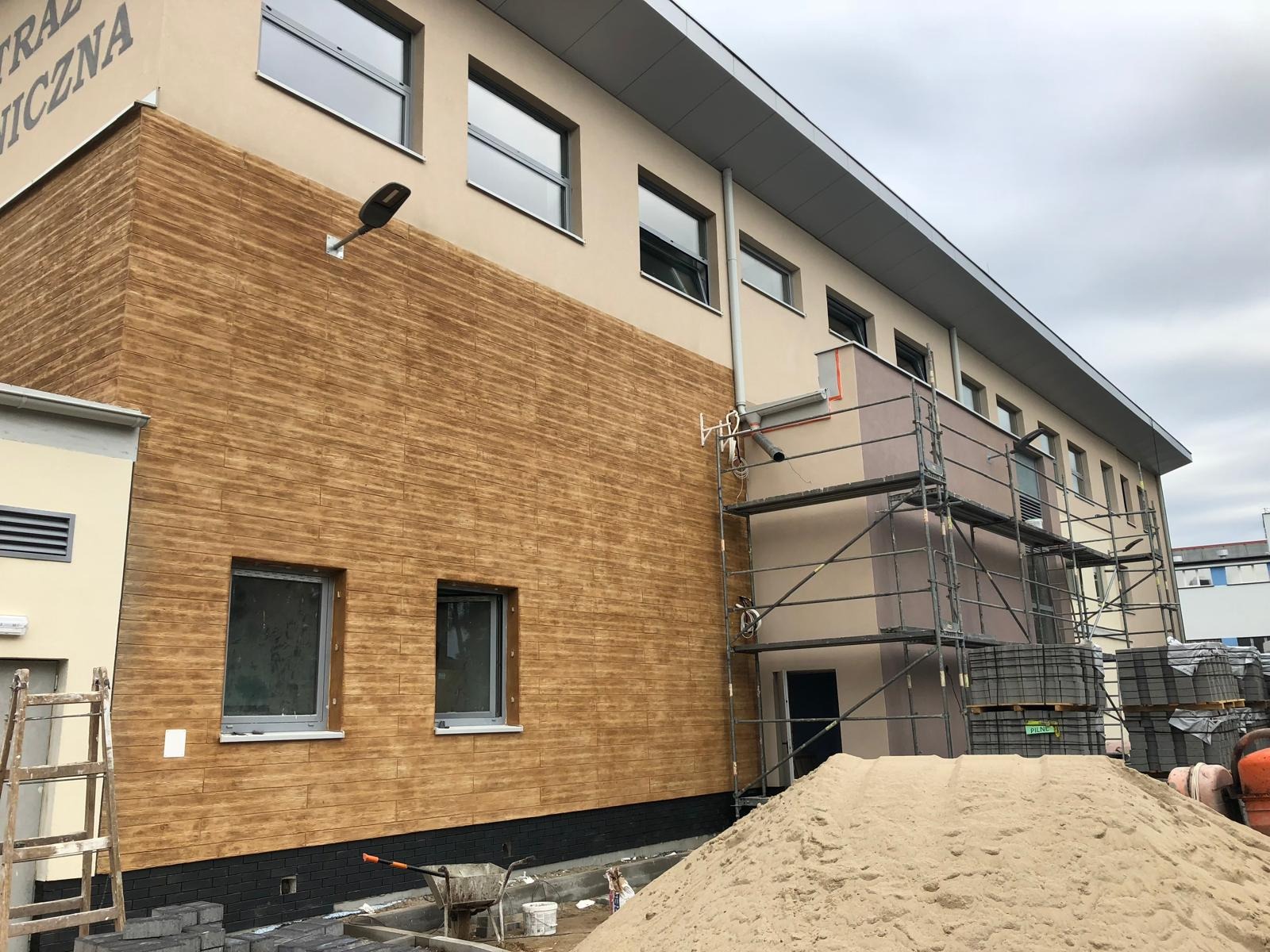 Budynek z elewacją imitującą drewno, rusztowania, piasek, i materiały budowlane wskazują na prace wykończeniowe.