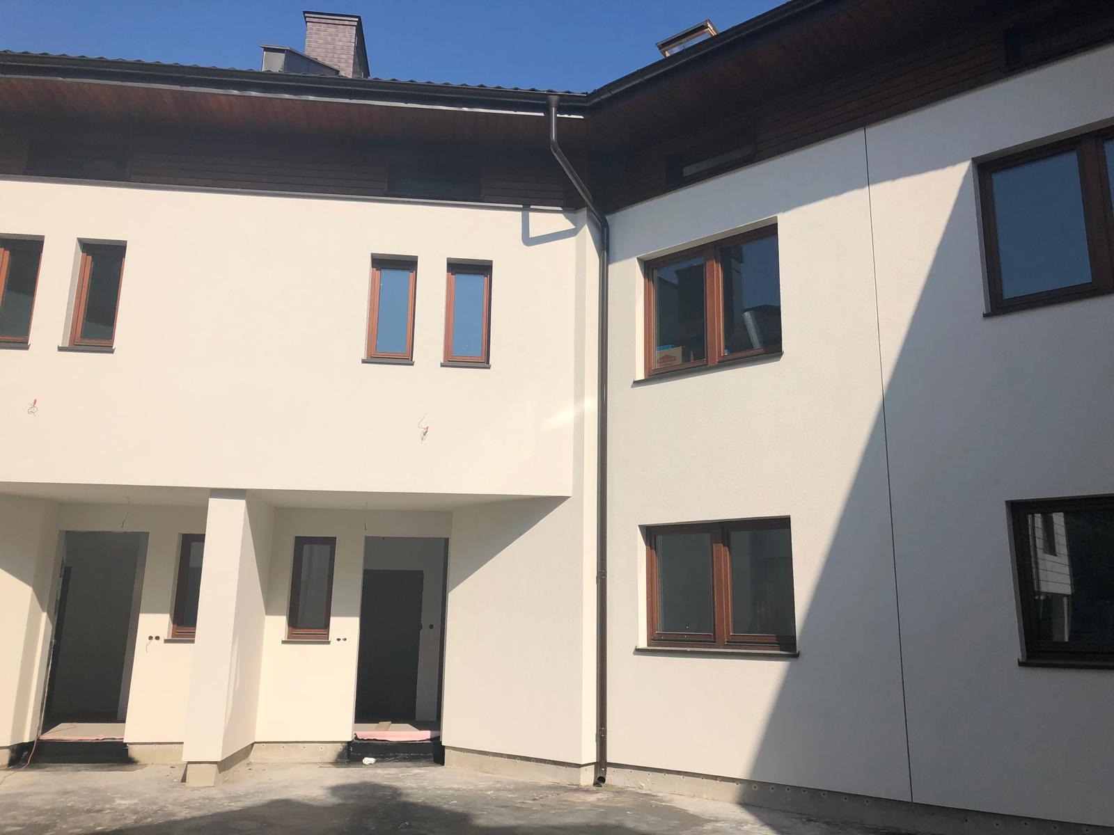 Nowoczesna elewacja szeregowca w jasnym odcieniu, z brązowymi oknami i orynnowaniem, widoczne niedokończone wejścia i fragment fundamentu z izolacją.