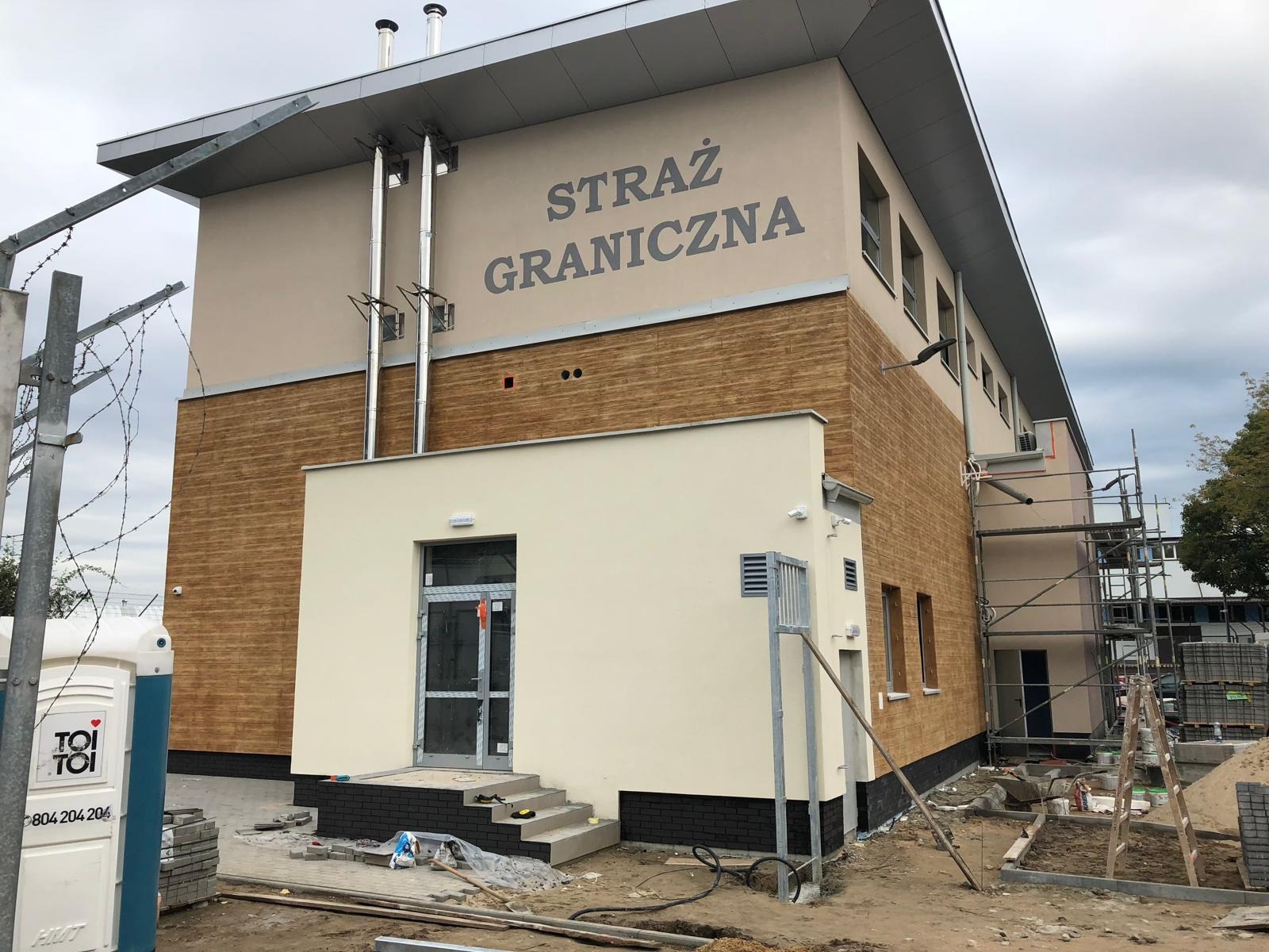 Budynek Straży Granicznej w trakcie prac wykończeniowych elewacji, widoczne rusztowania, przenośna toaleta i materiały budowlane.