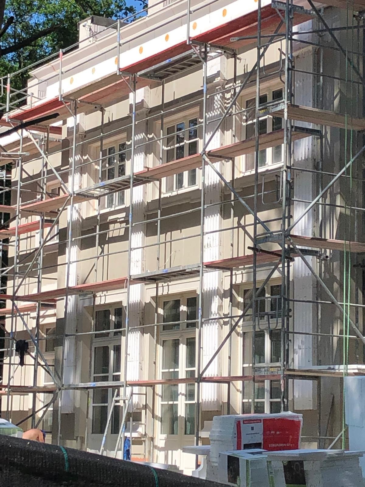 Elewacja budynku w trakcie prac, widoczne rusztowania, ocieplenie przy górnej krawędzi ściany i materiały budowlane u podstawy.