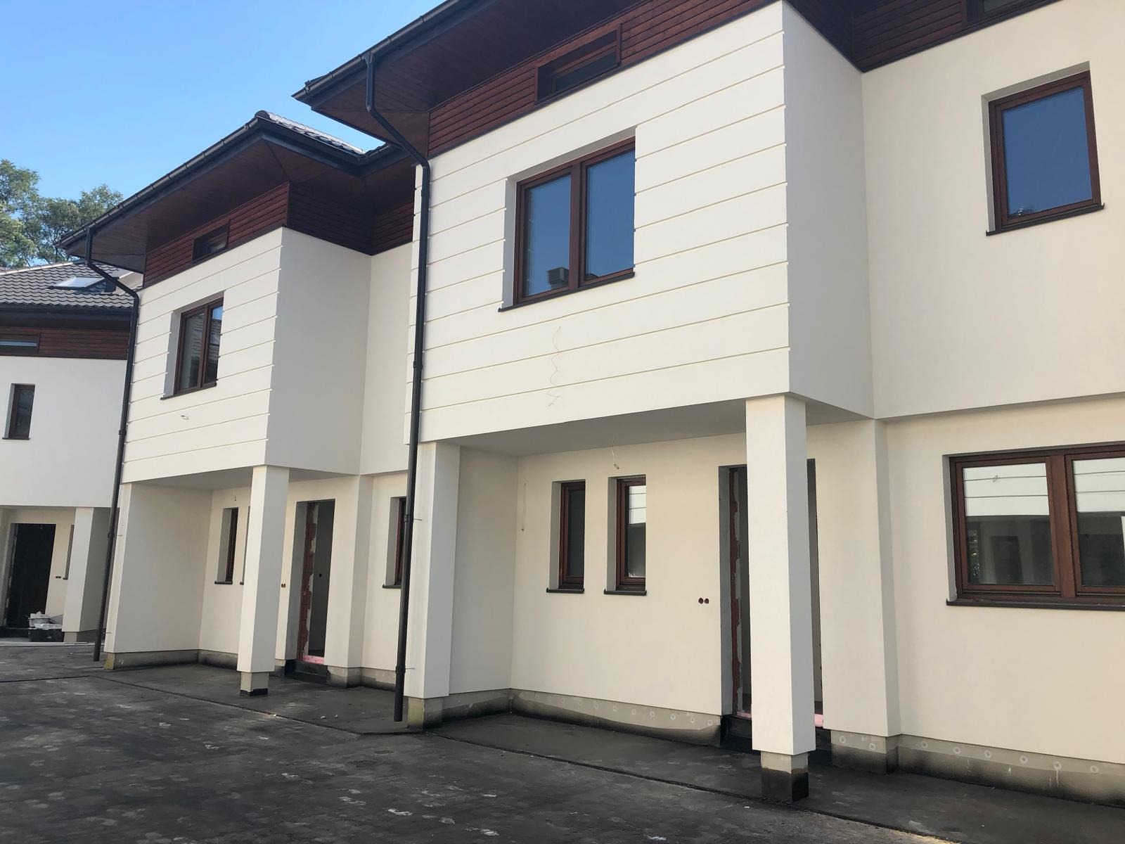 Nowoczesne szeregówki z białą elewacją, brązowymi ramami okiennymi i drewnianymi elementami wykończeniowymi dachu, w trakcie budowy lub tuż po zakończeniu prac.