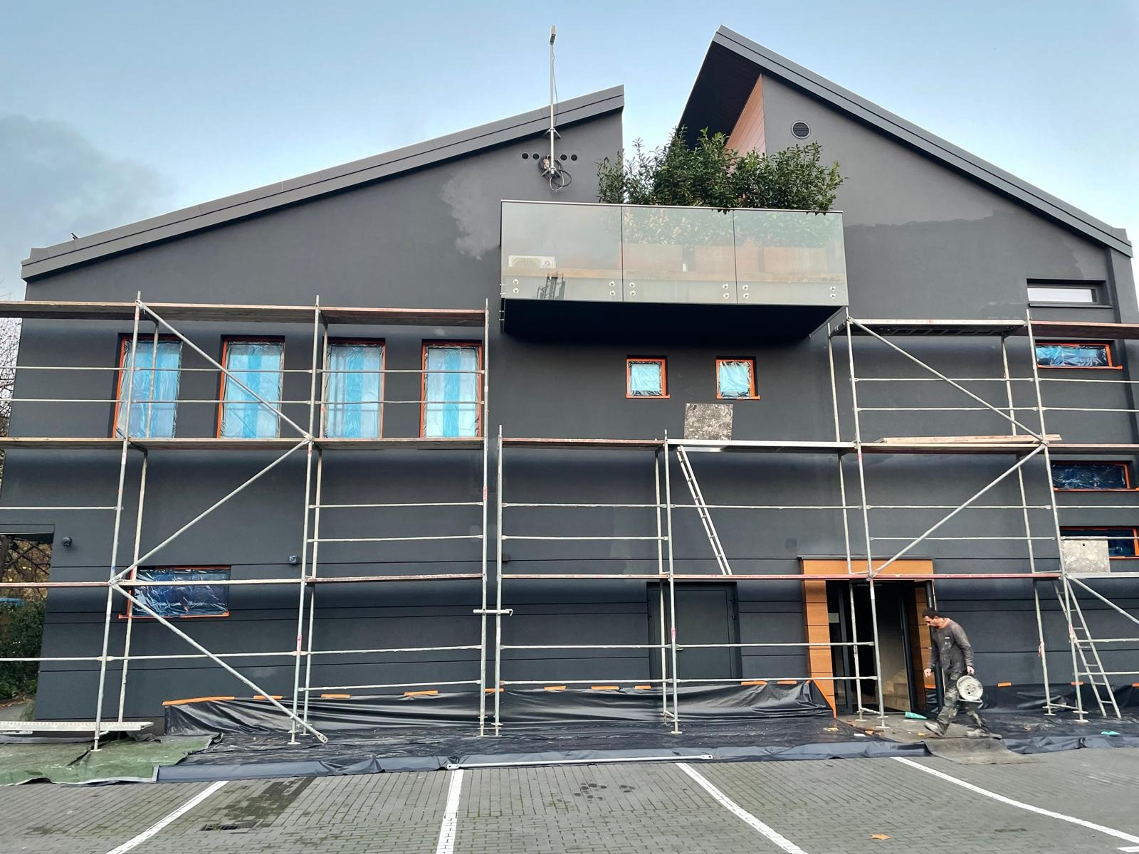 Malowanie elewacji nowoczesnego domu na czarno, z rusztowaniem, zabezpieczonymi oknami i pracownikiem wchodzącym do budynku.