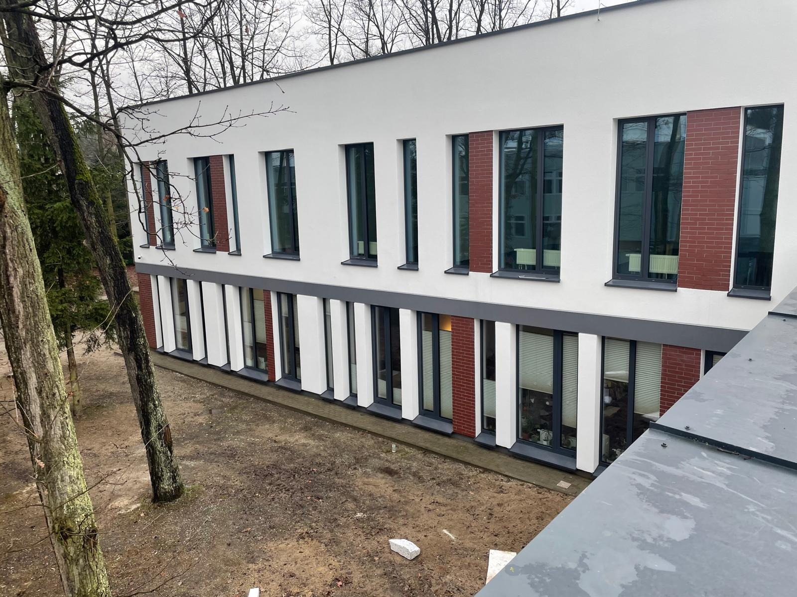 Dwukondygnacyjny budynek z białą elewacją i ceglanymi wstawkami wokół okien, widoczny z góry, z drzewami w tle i ziemią przed budynkiem.