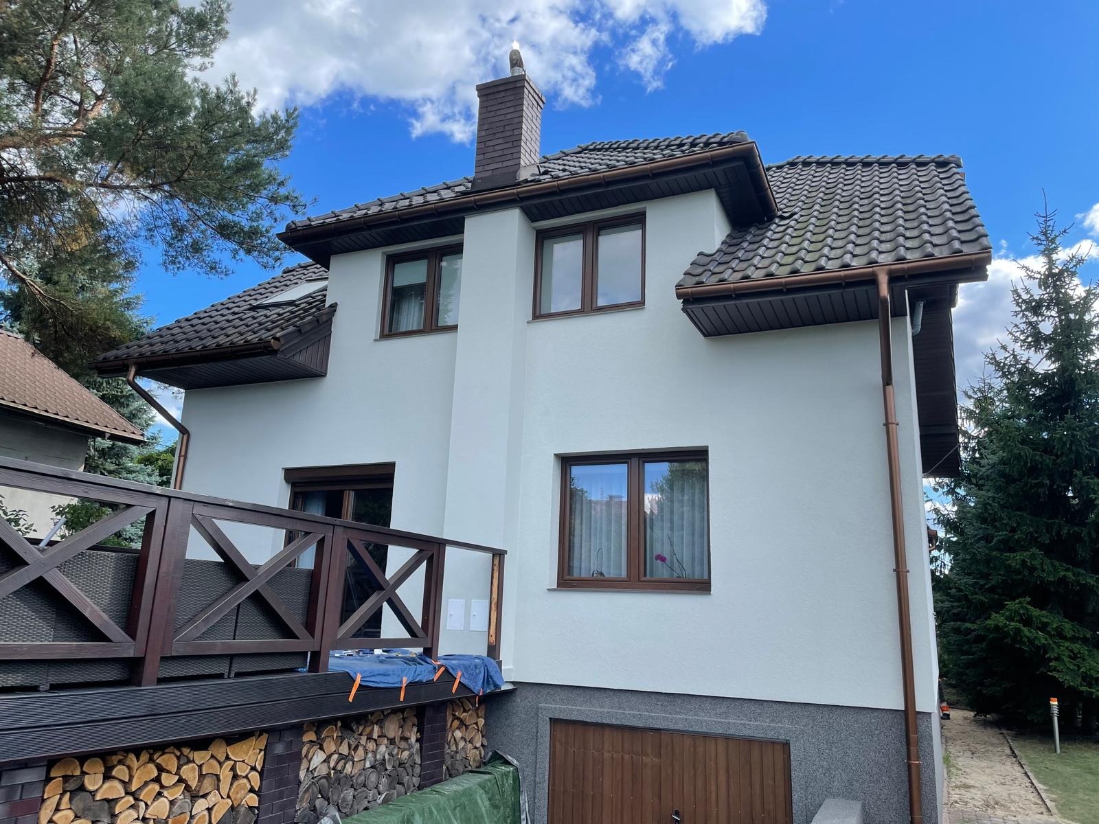 Nowoczesny dom z białą elewacją, brązowymi oknami i dachem z dachówki, widoczny z boku, z drewnianym balkonem i stosem drewna pod spodem.