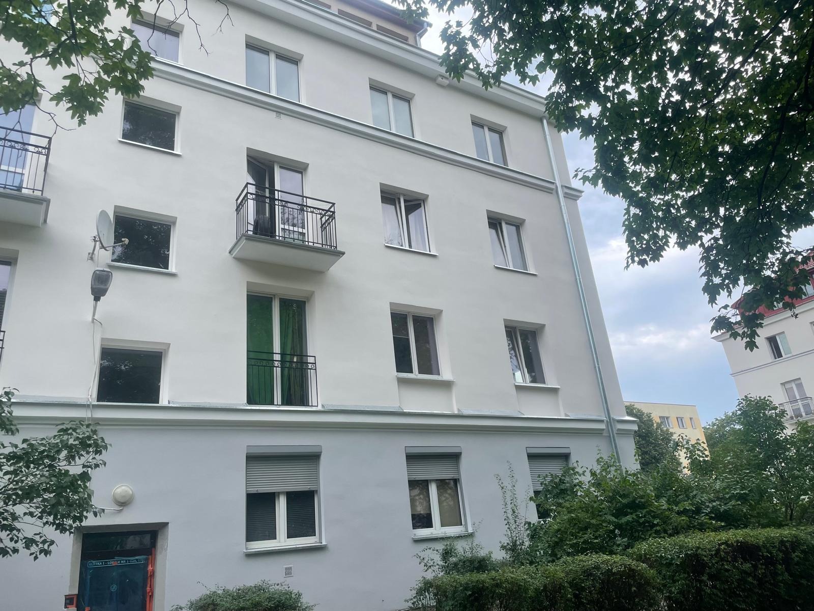 Odnowiona elewacja kamienicy z balkonami, widoczne okna z roletami i satelita na ścianie, zieleń w tle.