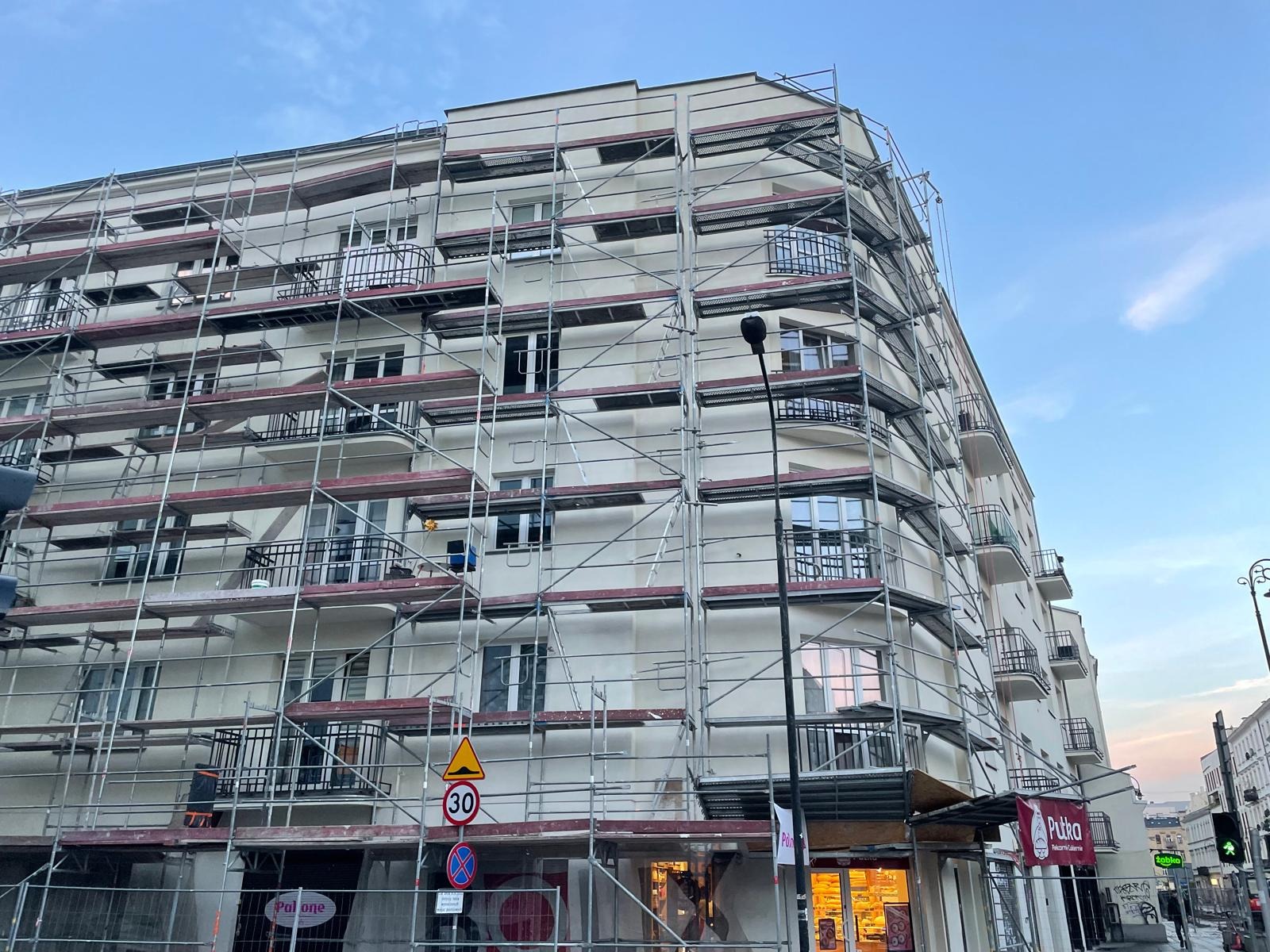 Budynek mieszkalny w trakcie prac elewacyjnych, otoczony rusztowaniem, z widocznymi znakami drogowymi i szyldem piekarni na parterze.