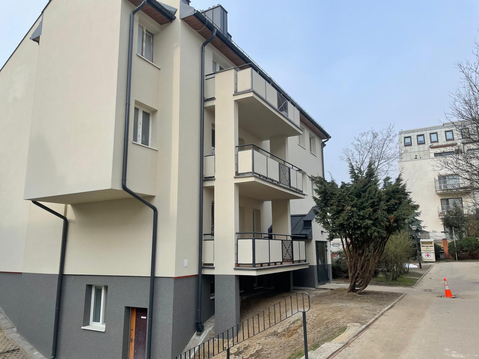 Nowoczesny budynek wielorodzinny z jasną elewacją, szarym cokołem, balkonami z metalowymi balustradami i rynnami, widok z dołu na tle drzewa i sąsiedniego budynku.