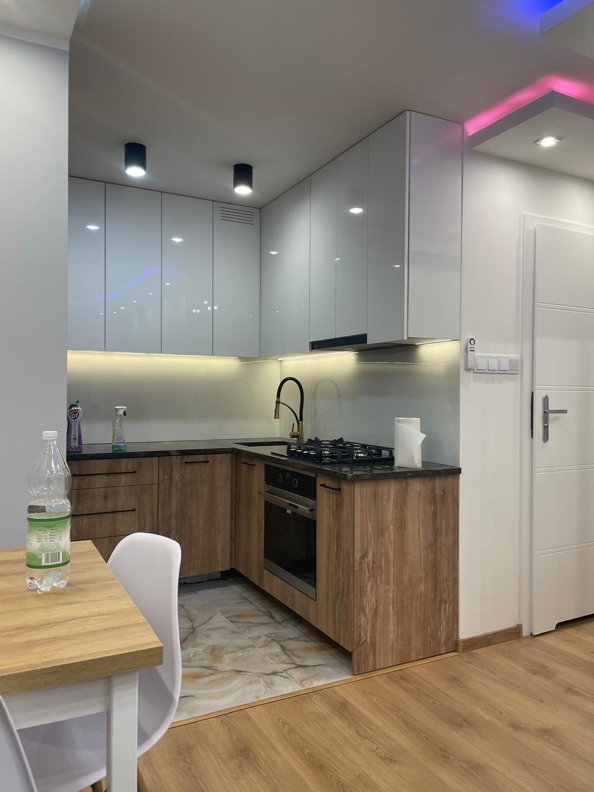 Nowoczesna kuchnia z białymi szafkami górnymi i drewnopodobnymi szafkami dolnymi, czarnym blatem, złotą baterią i podświetleniem LED, widok na fragment stołu z krzesłem.