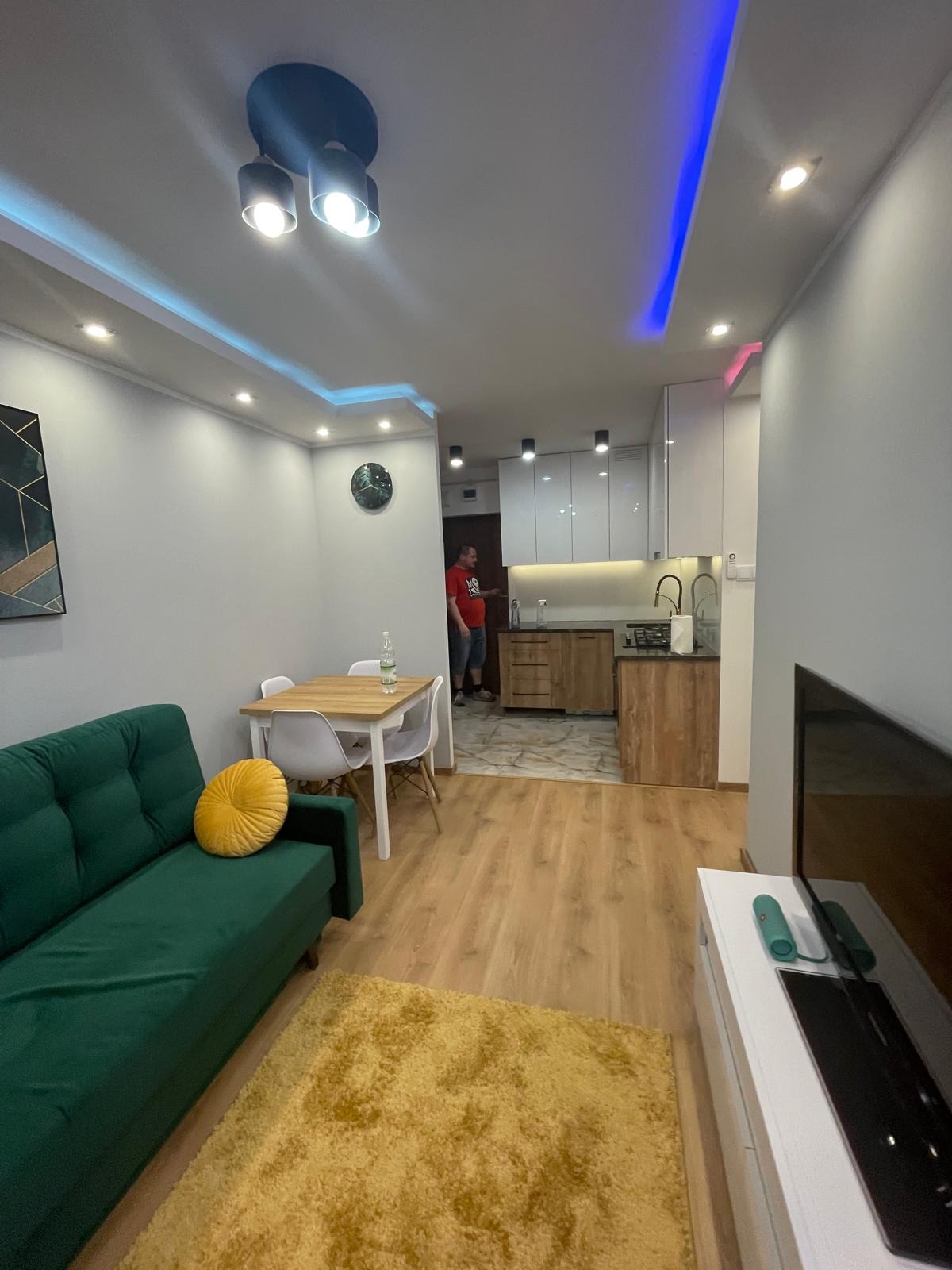Wnętrze nowocześnie urządzonego mieszkania z zieloną sofą, żółtym dywanem i oświetleniem LED, widok na otwartą kuchnię i osobę w tle.