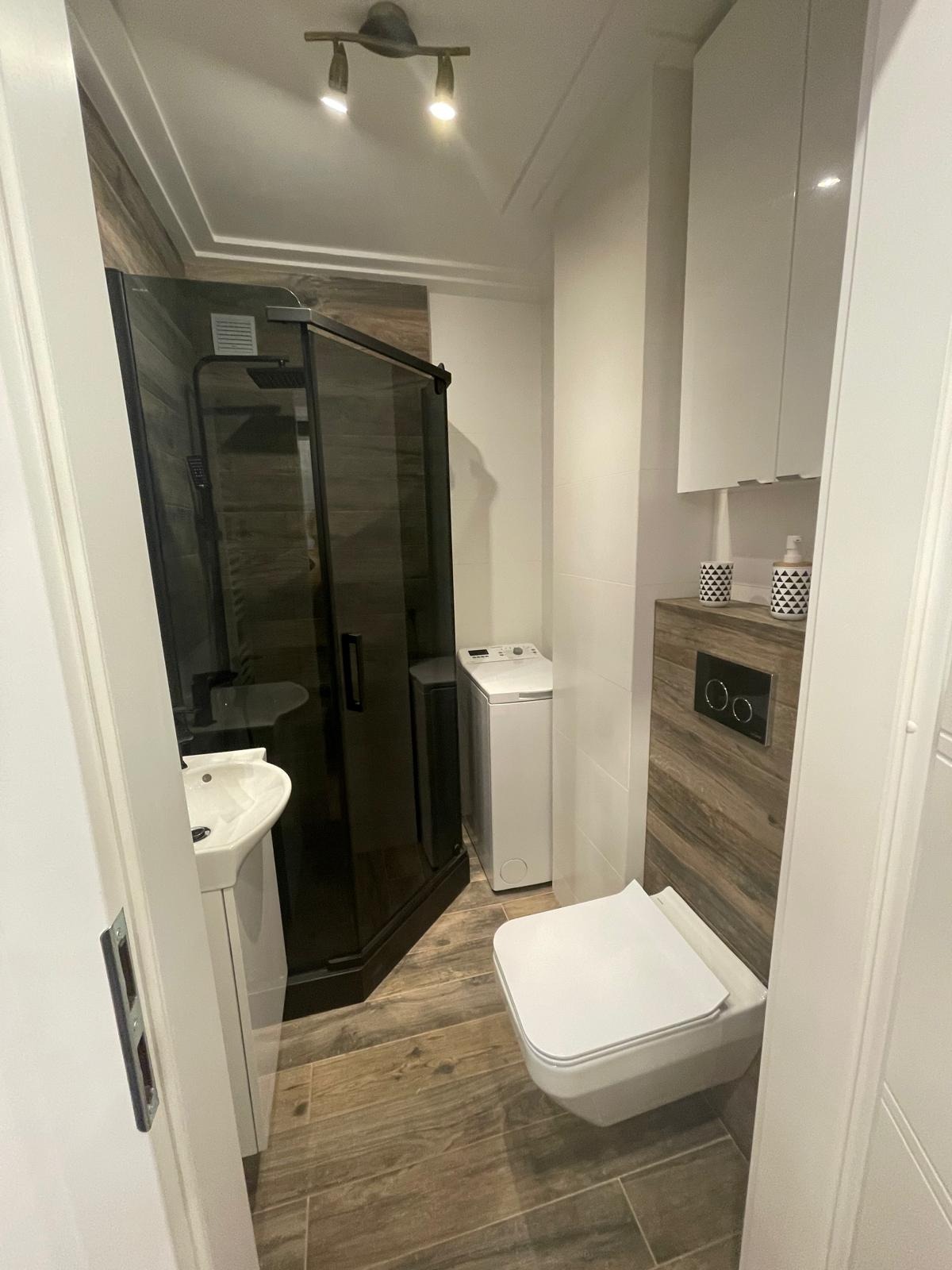 Nowoczesna łazienka z czarną kabiną prysznicową, umywalką na postumencie, podwieszaną toaletą i pralką, wykończona płytkami imitującymi drewno, oświetlona punktowymi lampami.