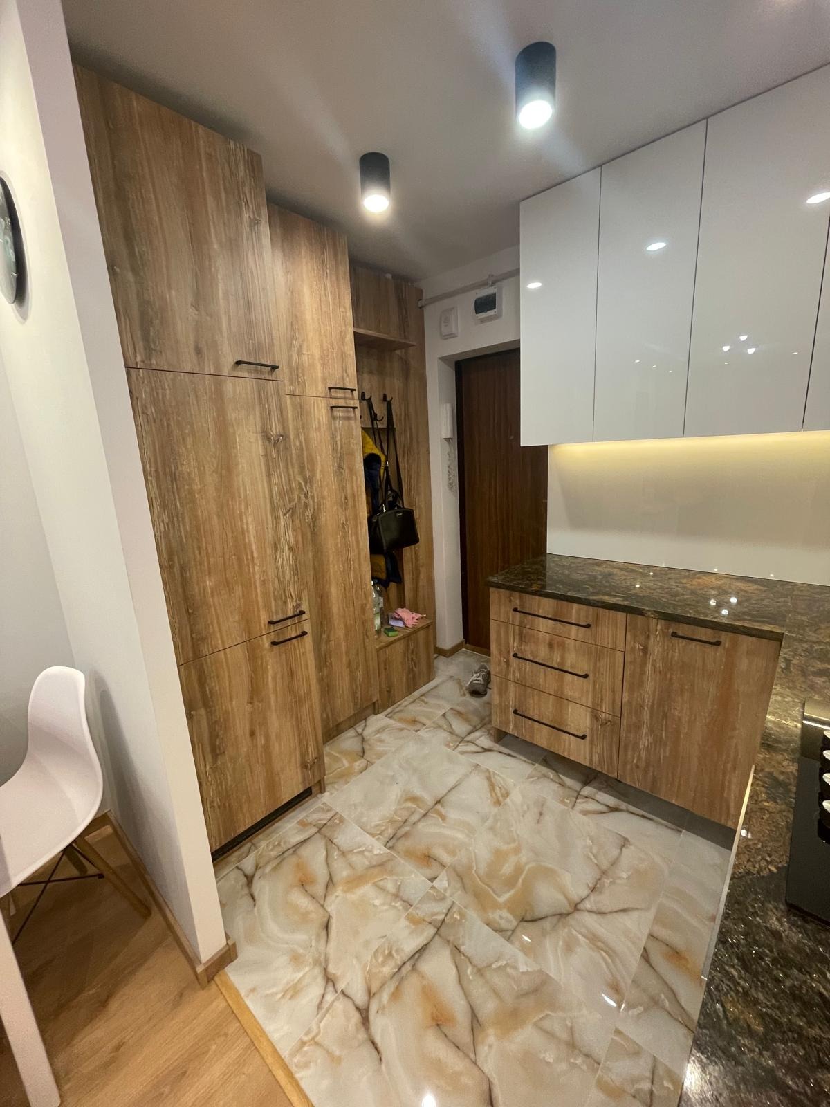 Wykończona kuchnia w Warszawie z drewnianymi szafkami i marmurową podłogą, widoczny fragment przedpokoju z wieszakami i szafką na buty.