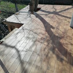 Nabłysk - Wykończony taras z płytek imitujących drewno, wbudowany w konstrukcję drzewa, z widocznymi cieniami gałęzi i fragmentem muru z cegły w tle.