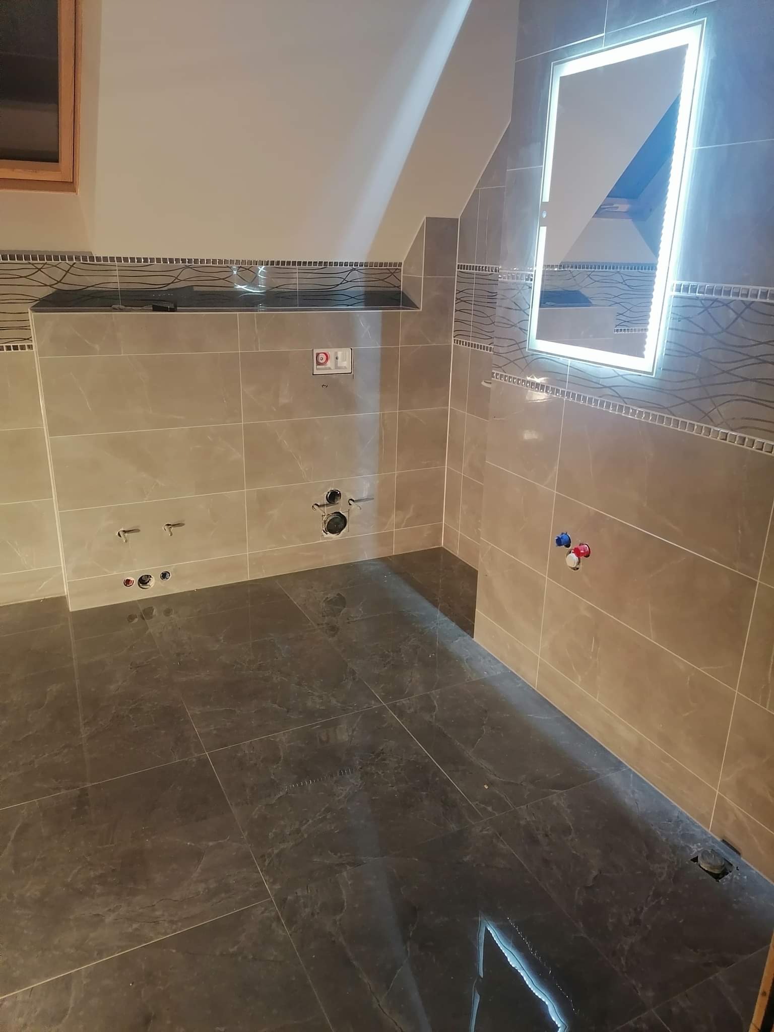 Przygotowana do montażu armatury łazienka z beżowymi płytkami ściennymi, ciemną podłogą z płytek imitujących marmur i podświetlanym lustrem LED.