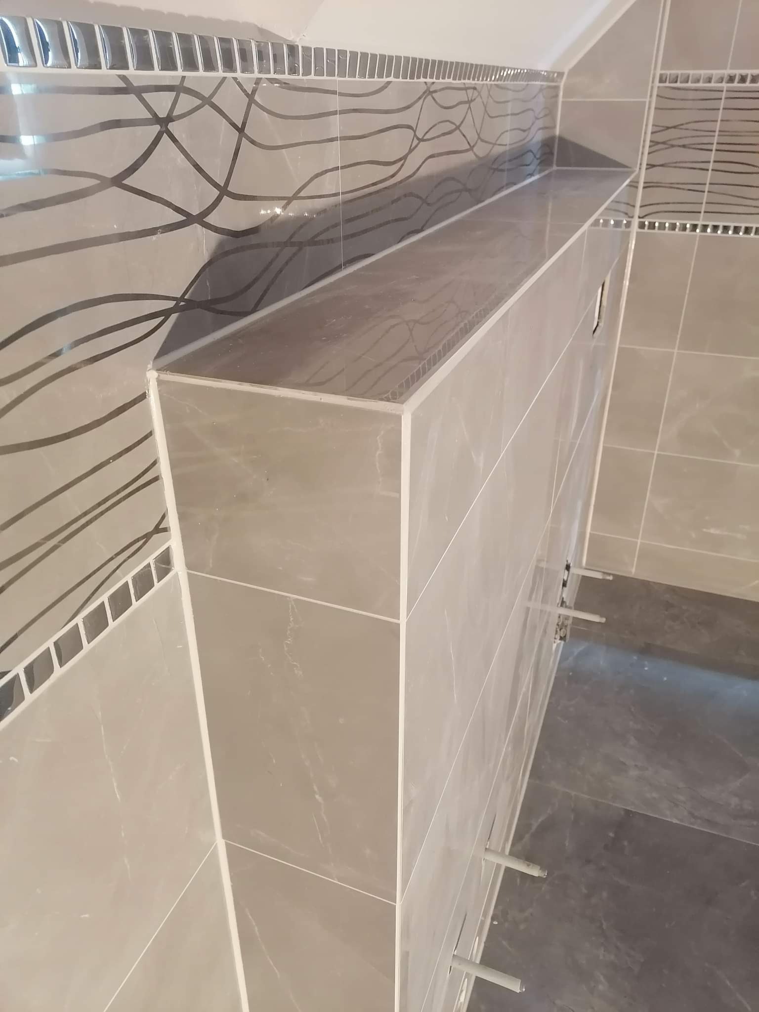 Półka z szarych płytek w łazience, dekoracyjne płytki mozaikowe na ścianie, widoczne plastikowe kołki montażowe.