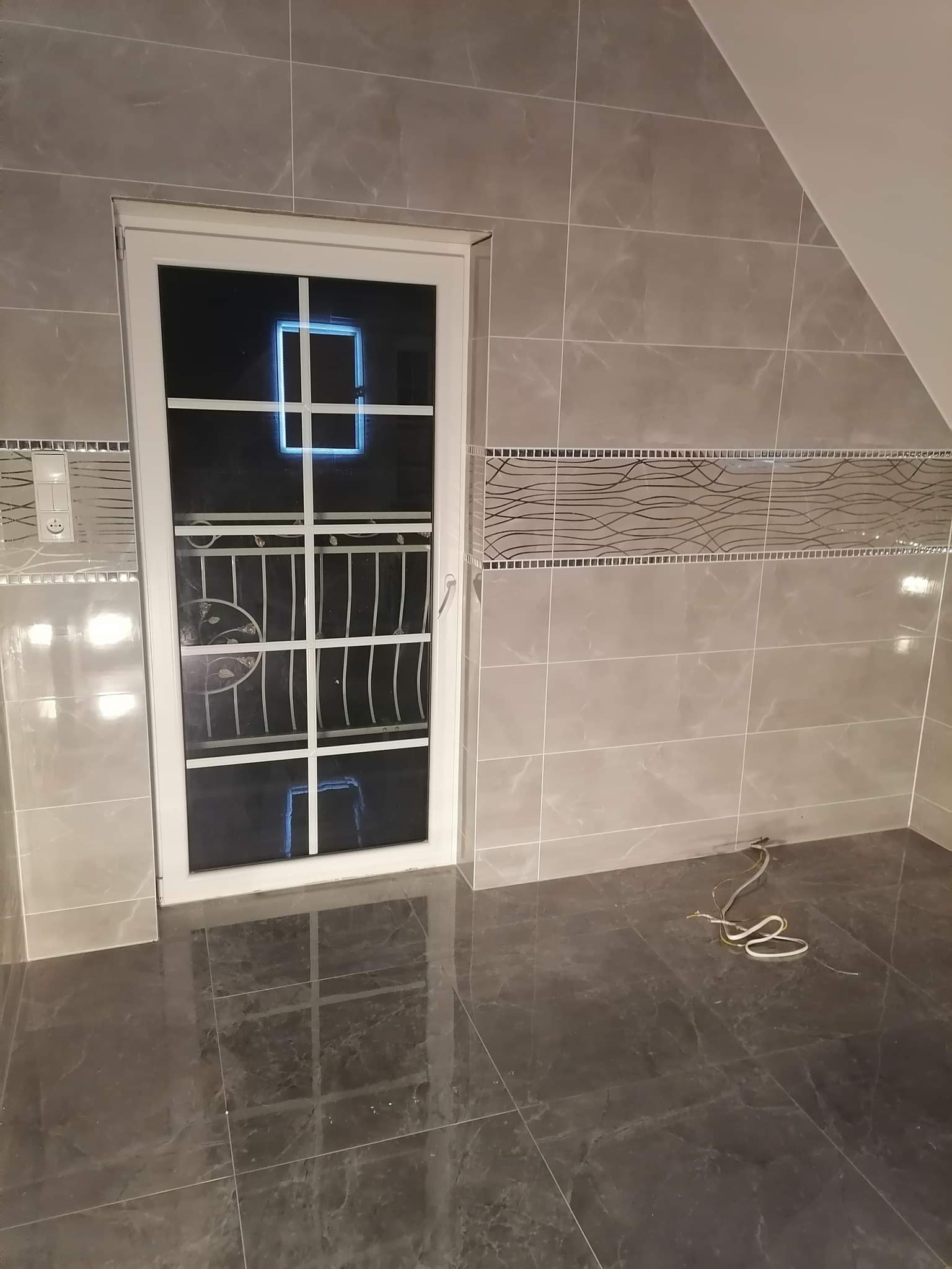 Wykończona łazienka na poddaszu z szarymi płytkami imitującymi marmur na ścianach i podłodze, białe okno z kratką i dekoracyjny pas płytek na ścianie