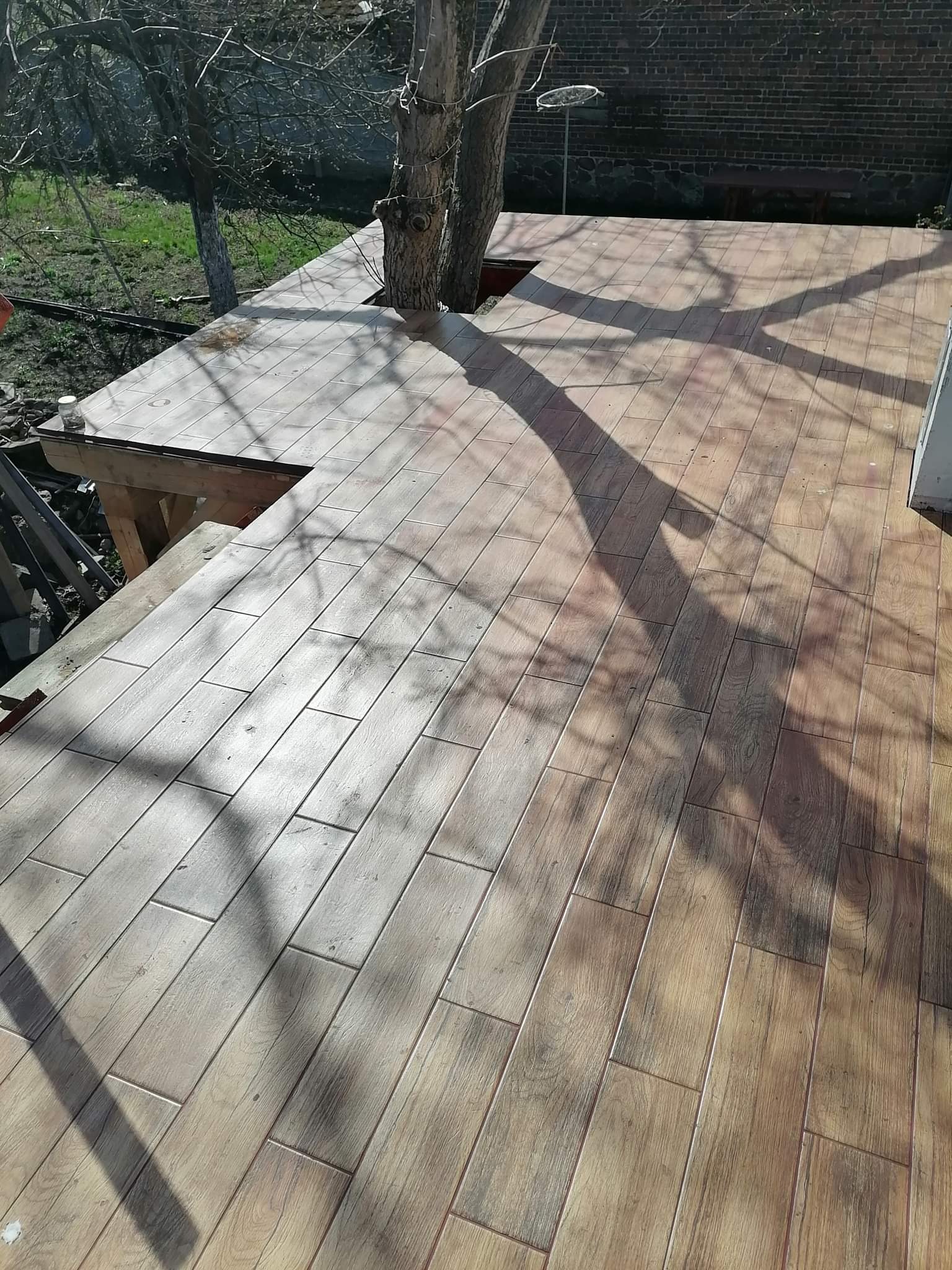 Wykończony taras z płytek imitujących drewno, wbudowany w konstrukcję drzewa, z widocznymi cieniami gałęzi i fragmentem muru z cegły w tle.