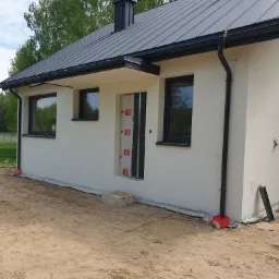 Ocieplony dom jednorodzinny z czarną stolarką okienną i grafitowym dachem w Jadowie, widok z zewnątrz na elewację w trakcie prac wykończeniowych.