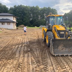 Instalacje w Twoim Domu - Żółta koparko-ładowarka JCB na placu budowy domu jednorodzinnego, widoczny mężczyzna w tle, teren wyrównany z odciśniętymi śladami opon.