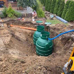 Instalacje w Twoim Domu - Dwa zielone zbiorniki w wykopie ziemnym, pomarańczowa rura i niebieski wąż, teren budowy z roślinnością w tle. Widoczny pracownik i sprzęt budowlany.