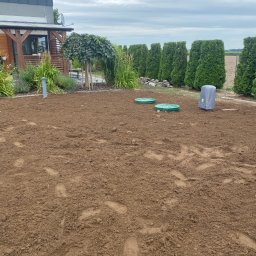 Instalacje w Twoim Domu - Przygotowany teren pod przydomową oczyszczalnię ścieków z widocznymi studzienkami i zbiornikiem w szarej obudowie. W tle rząd tuj i dom.