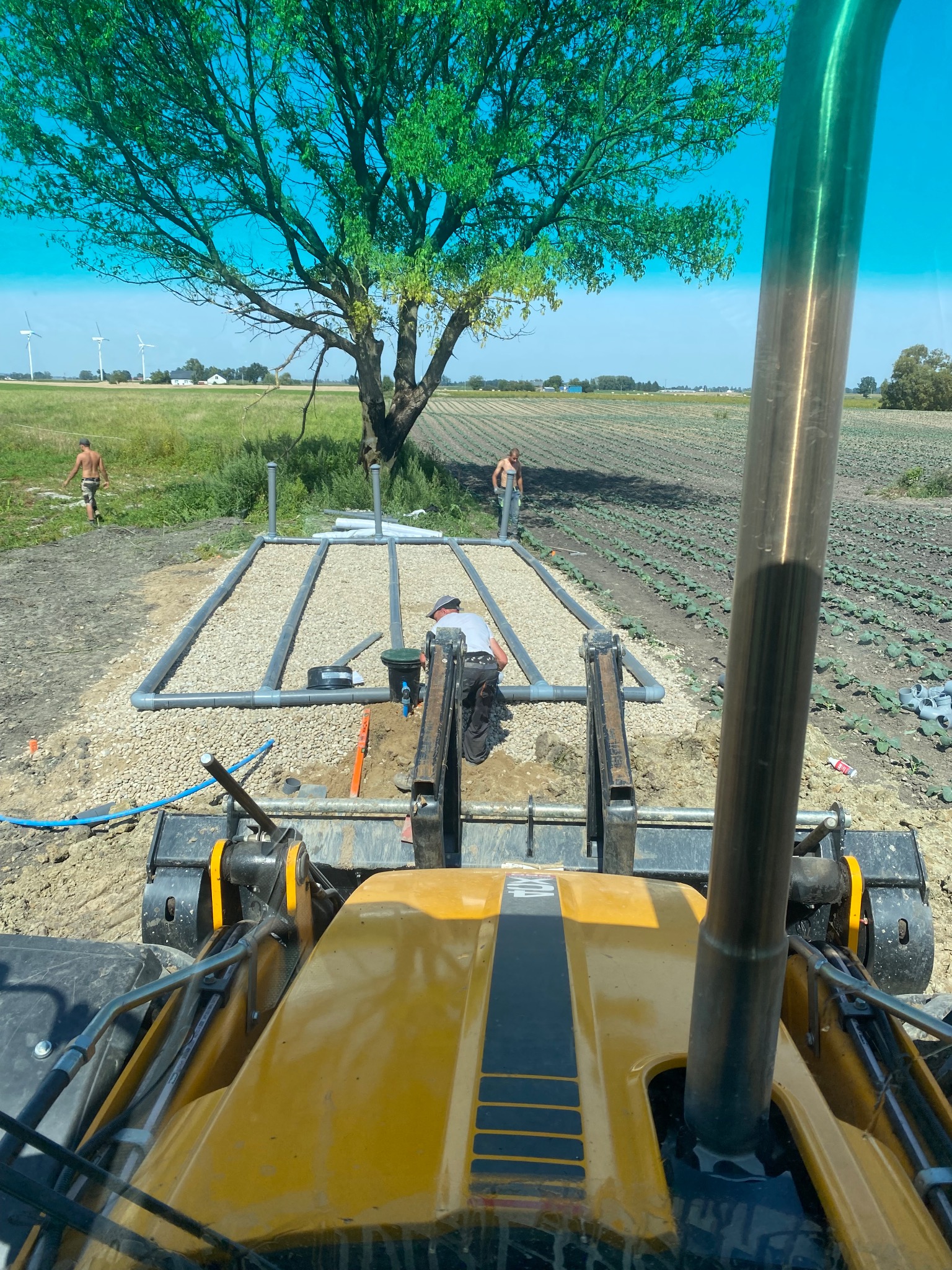 Montaż drenażu rozsączającego z perspektywy operatora koparki. Widoczne rury drenażowe ułożone w gruncie, żwir, oraz pracownicy na tle pola i wiatraków.