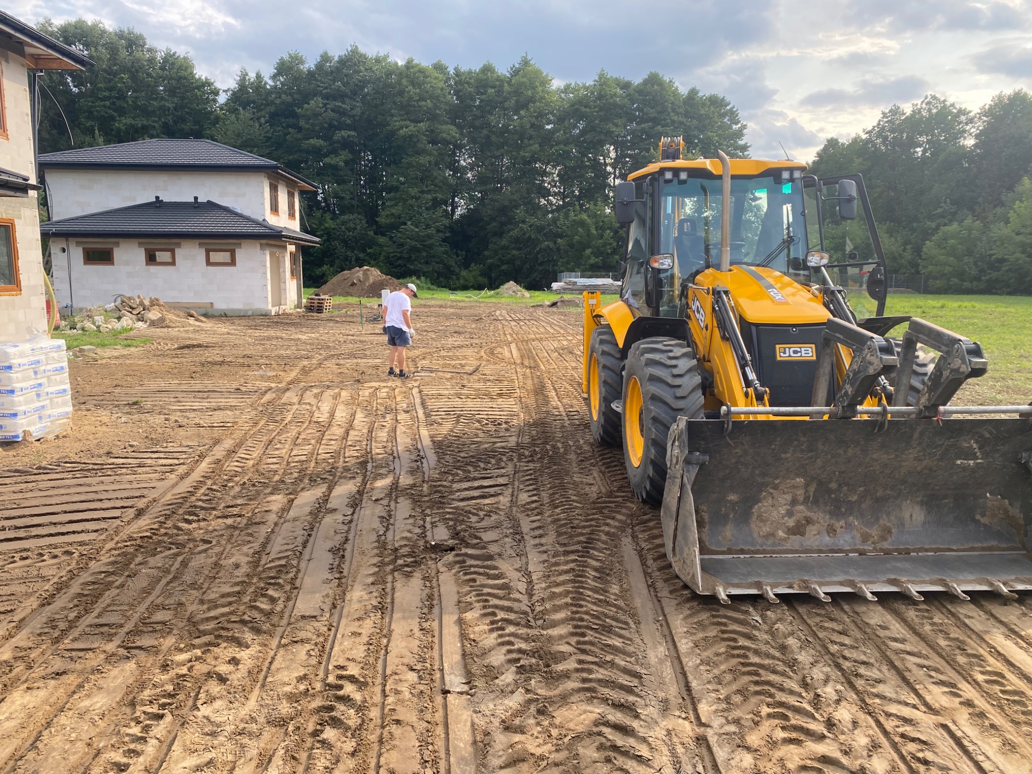Żółta koparko-ładowarka JCB na placu budowy domu jednorodzinnego, widoczny mężczyzna w tle, teren wyrównany z odciśniętymi śladami opon.